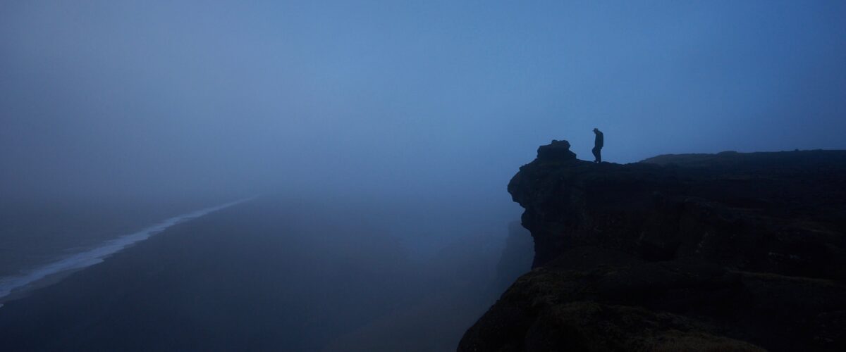 a person standing on a cliff, overlooking the ocean with a dark blue, moody sky